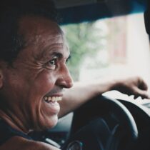Esmer kavruk bir adam sürücü koltuğunda yanındaki arkadaşına bakarak gülüyor.