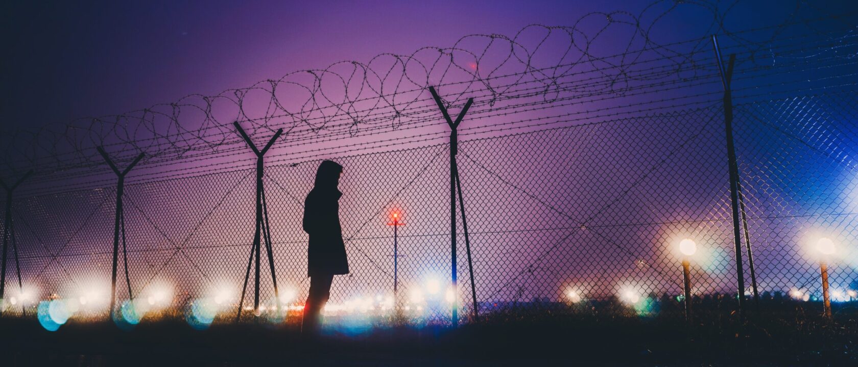 Yüksek hapishane çitlerinin ardından şehre bakan bir insan silüeti.
